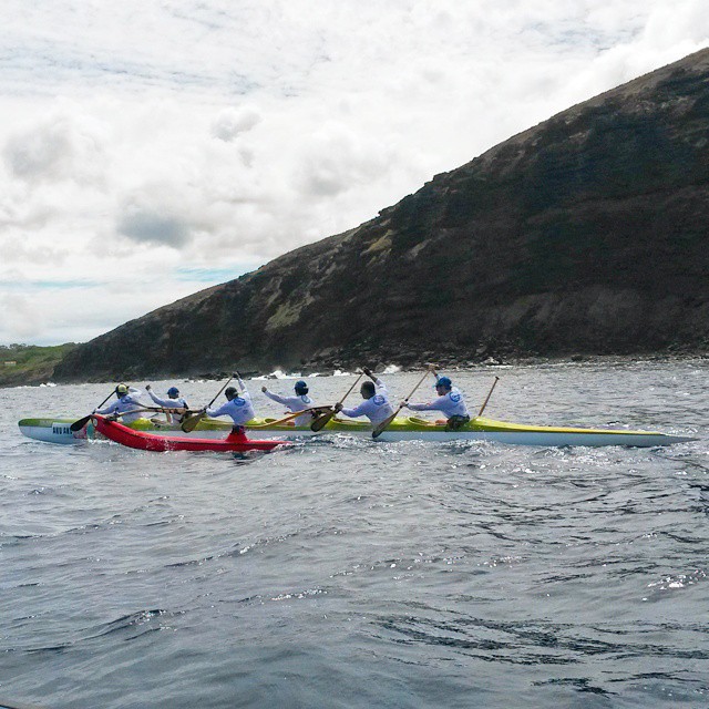 Un equipo de seis personas con camisas blancas rema en una canoa en el océano cerca de una costa rocosa. El agua está ligeramente agitada y el cielo está nublado. La canoa es multicolor con secciones amarillas, rojas y verdes.