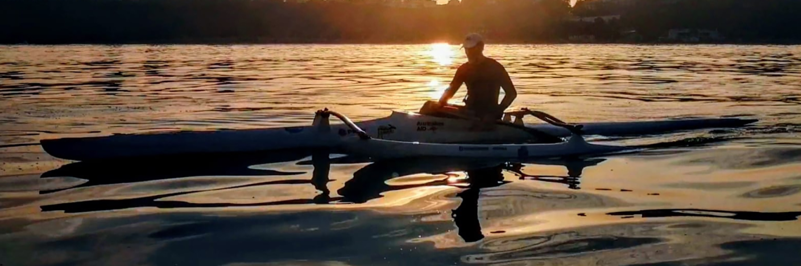 Una persona rema en kayak por aguas tranquilas al atardecer, creando suaves ondulaciones en la superficie reflectante. El sol proyecta un cálido resplandor que recorta la silueta del kayakista contra un fondo de colinas distantes.