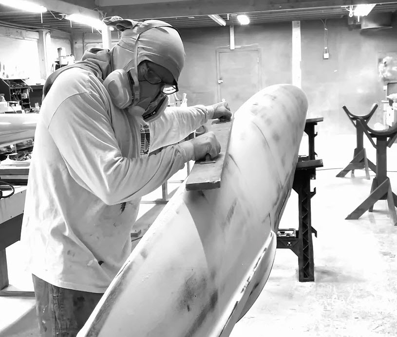 Una persona con equipo de protección lija un objeto grande y liso en un taller. El entorno es industrial, con soportes y herramientas a su alrededor. La imagen es en blanco y negro, lo que resalta los detalles del espacio de trabajo.