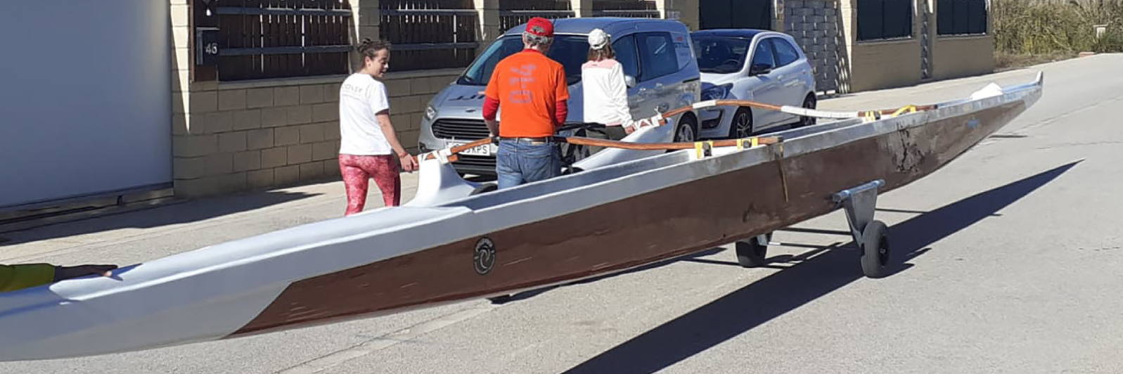 Tres personas, dos de ellas con gorras, maniobran una gran embarcación parecida a una canoa sobre un carro con ruedas por una calle. Al fondo se ven una furgoneta blanca y un coche aparcado en un día soleado.