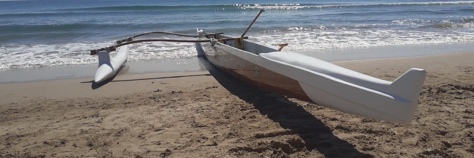 Una canoa con estabilizadores descansa sobre una playa de arena con suaves olas rompiendo contra la orilla. Se ven el casco principal y el estabilizador, y el océano se extiende hasta el horizonte bajo un cielo despejado.